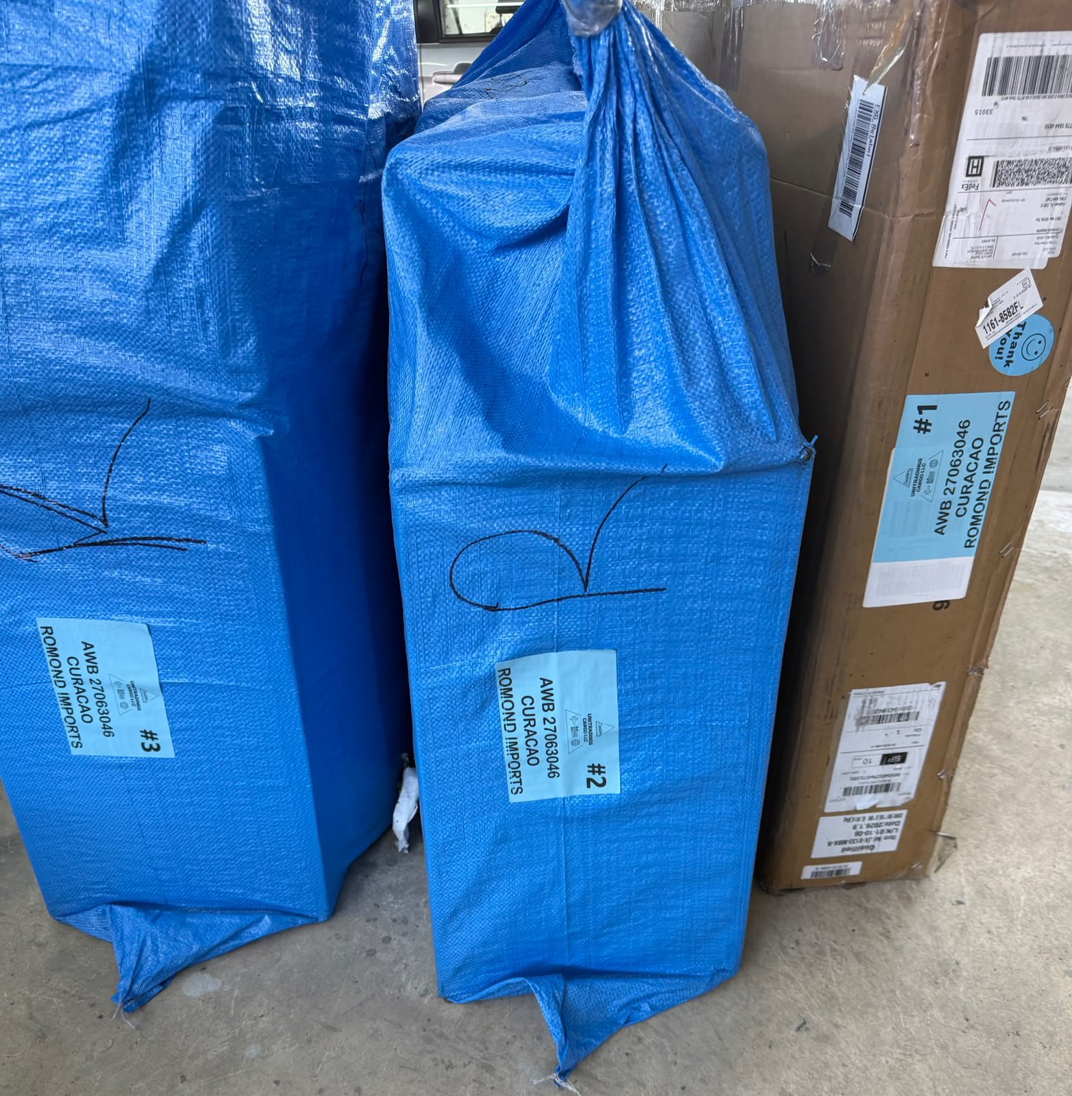 Packages wrapped in blue tarp, labeled AWB, Curaçao, Romond Imports, ready to fly